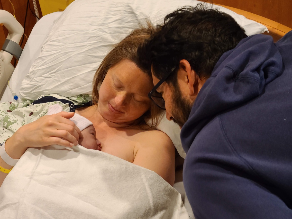 A woman with a newborn baby laying on her chest and a man is leaning in smiling